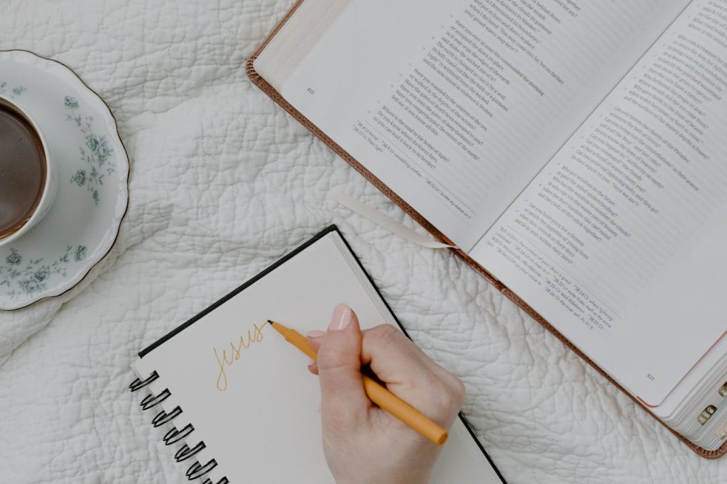 pexels-photo-8383482 A hand writing in a notebook next to an open Bible and a cup of coffee on a quilted blanket.